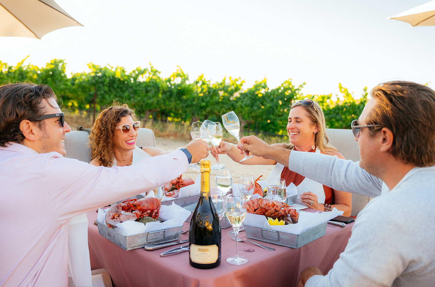 Outdoor table by the vineyard with guests toasting flutes of sparkling wine and plates of lobster
